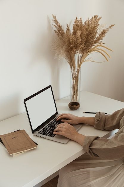 Person typing on laptop at minimalist desk with pampas grass vase and notebook.