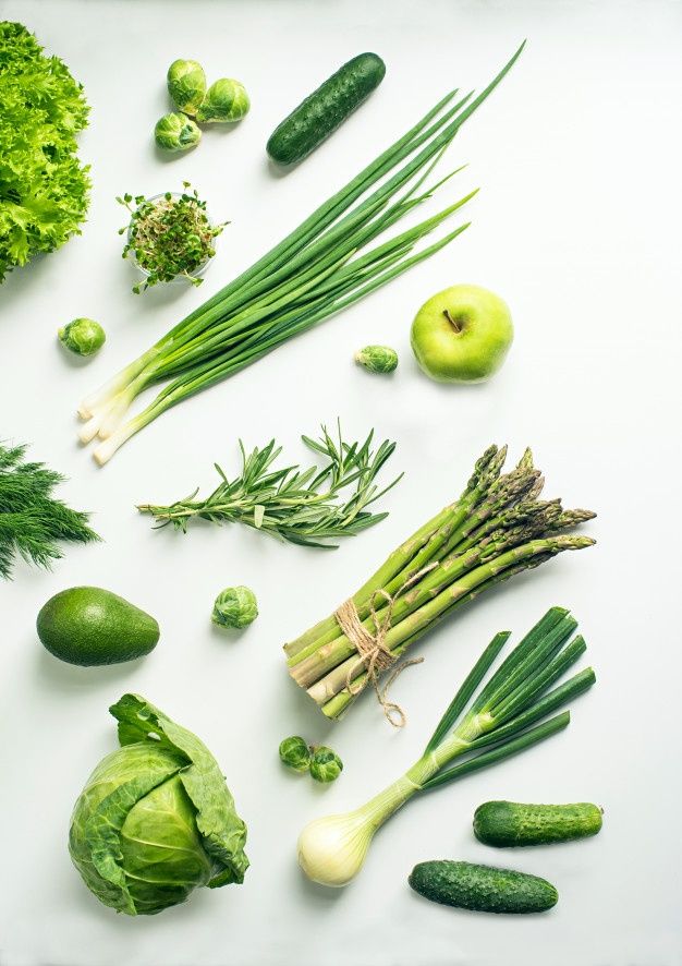 Assorted fresh green vegetables including asparagus, cabbage, apple, and cucumbers on a light background.