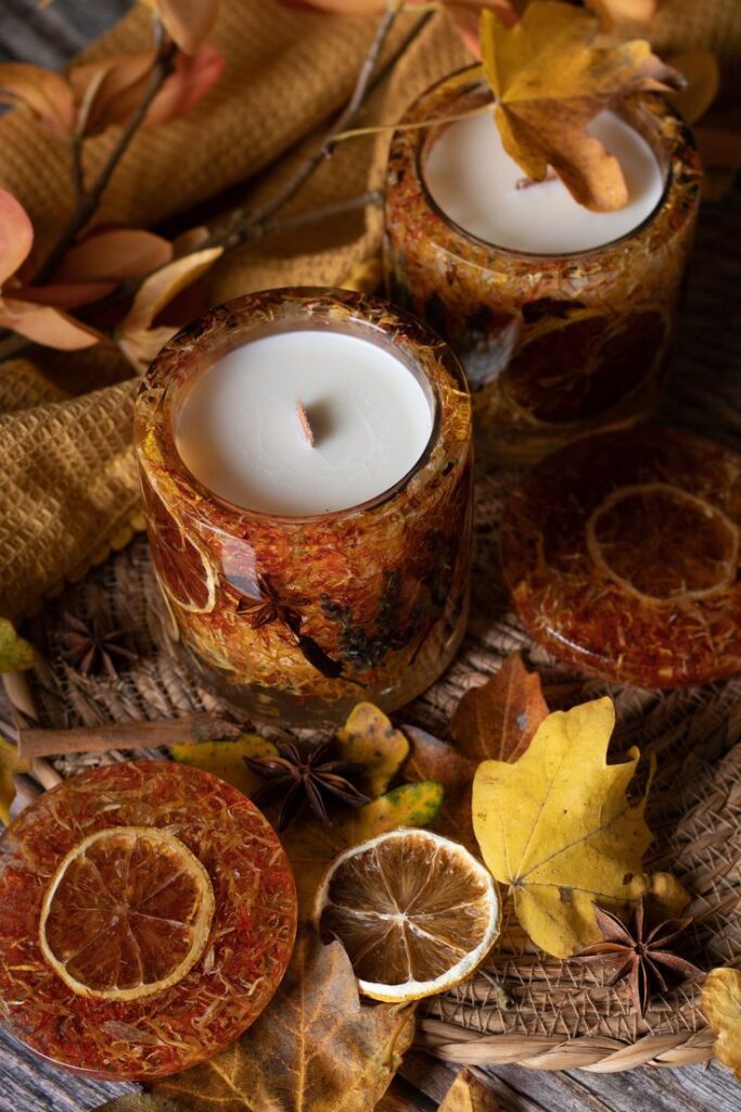 Autumn-themed candles with dried oranges and leaves for cozy home decor, placed on woven mat. Perfect for fall ambiance.