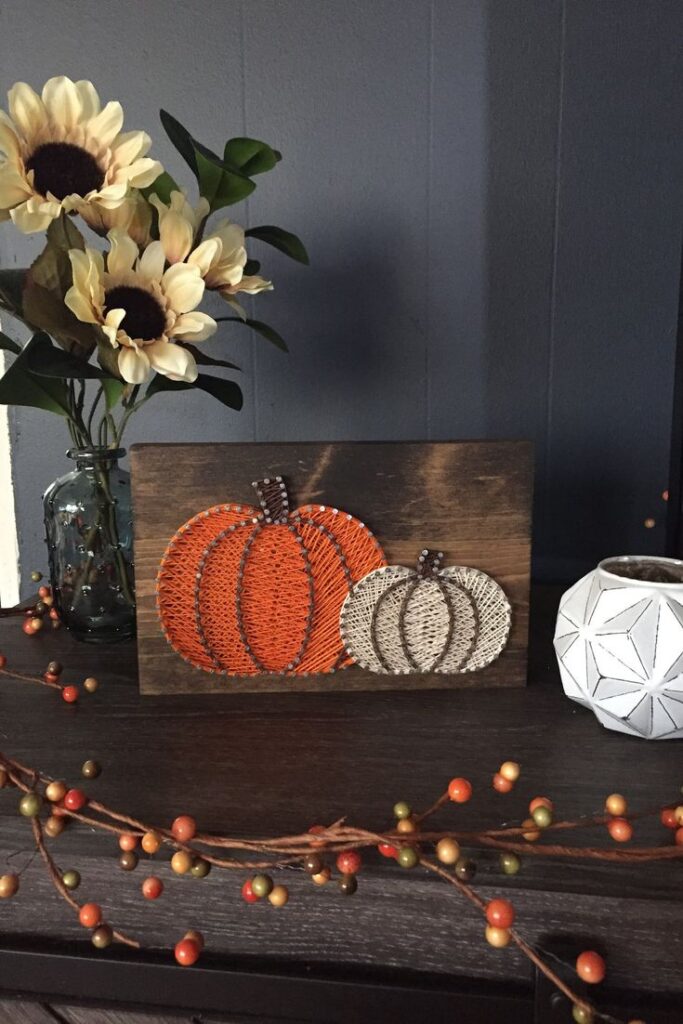 Rustic fall decor with sunflower vase and pumpkin string art on a wooden board. Cozy autumn home accents.