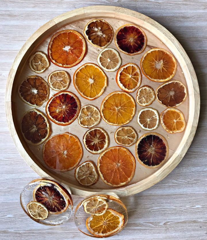 Dried citrus slices in a wooden tray with two filled glasses on a wooden table surface.