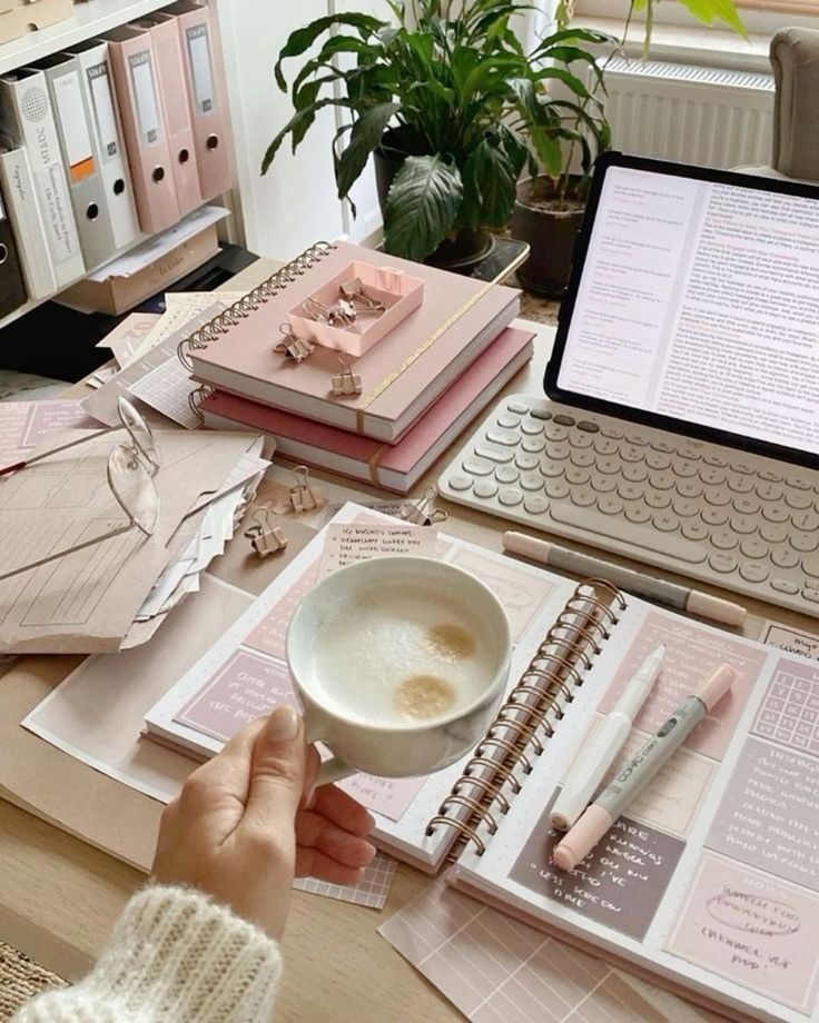 Cozy workspace with notebooks, a laptop, and a cup of coffee, surrounded by plants and stationery in pastel pink tones.