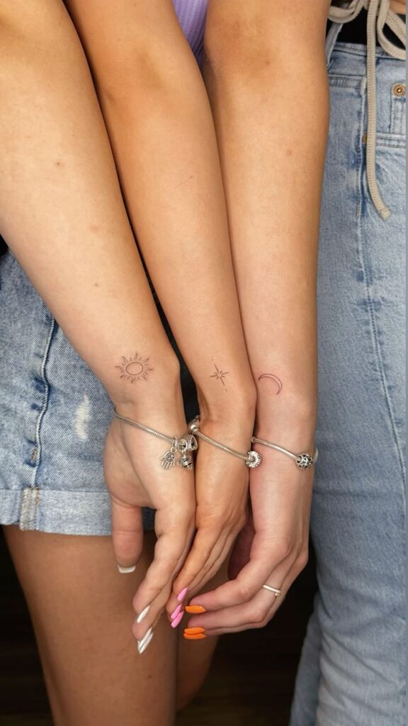 Matching arm tattoos of sun, star, and moon with charm bracelets on intertwined arms in denim shorts.