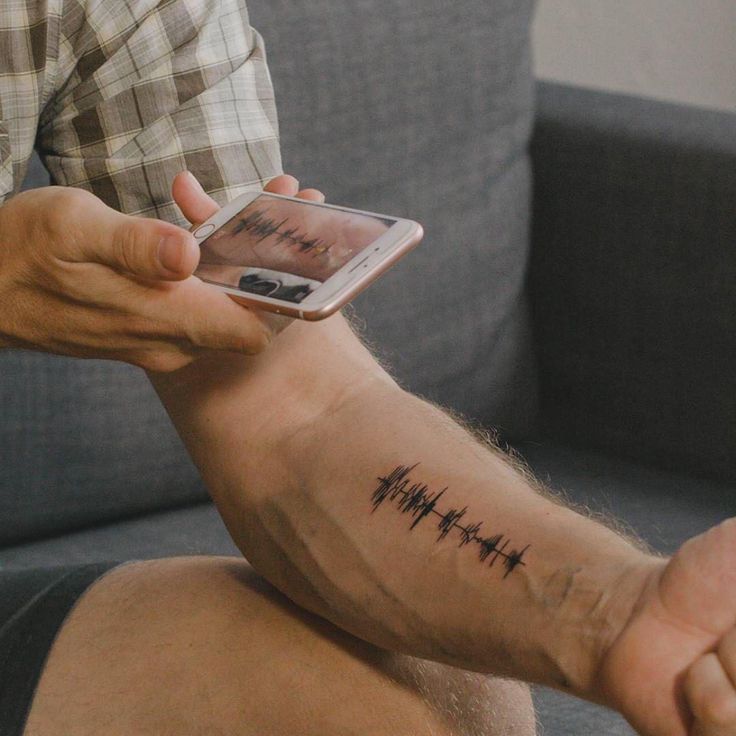 Person photographing forearm soundwave tattoo with smartphone.