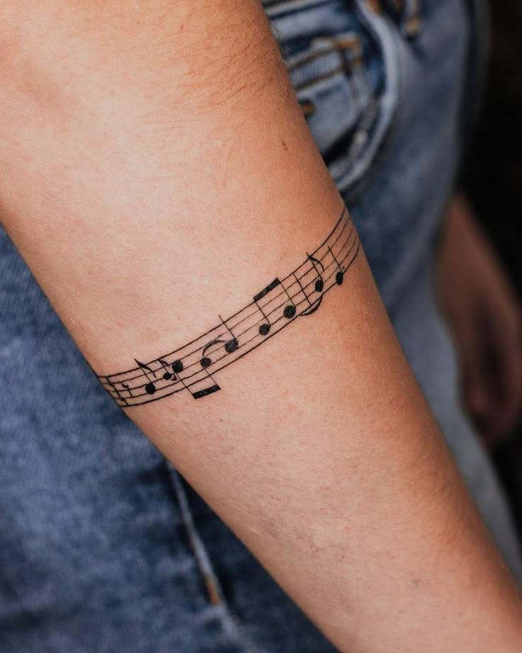 Musical notes tattoo wraps around forearm; creative body art in black ink on denim background.