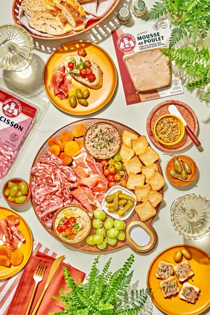 Charcuterie board with meats, cheeses, olives, bread, and spreads, artfully arranged on a table with garnishes.