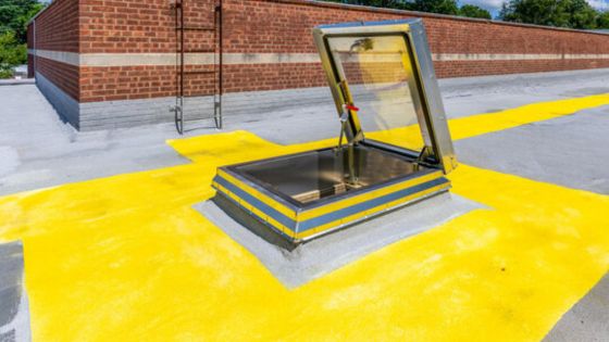 Rooftop access hatch with open door, highlighted by vibrant yellow safety markings, brick wall and ladder in background.