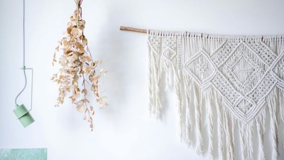 Macramé wall hanging with geometric patterns beside dried foliage on minimalist white background.