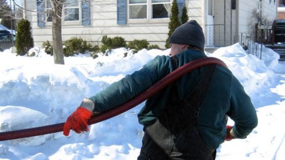 Worker delivering oil in winter, holding a hose, surrounded by snow in front of a house.