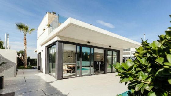 Modern luxury villa with glass walls, palm trees, and a spacious patio under a clear blue sky.