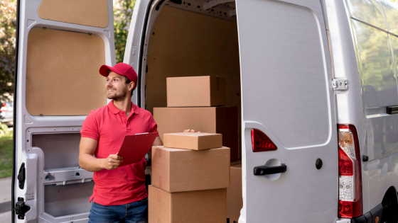 Delivery worker in red uniform unloading cardboard boxes from a van, holding a clipboard and smiling.