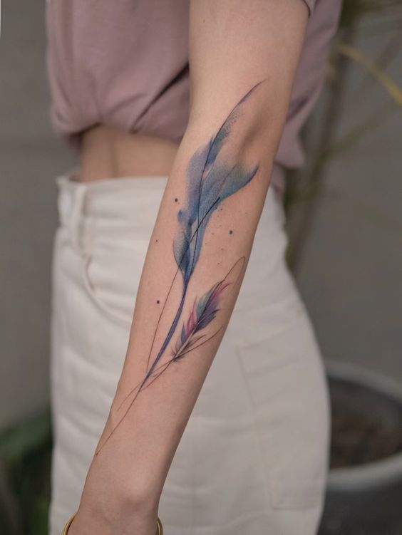 Elegant watercolor feather tattoo on forearm with soft blue and purple hues, showcasing artistic body art.