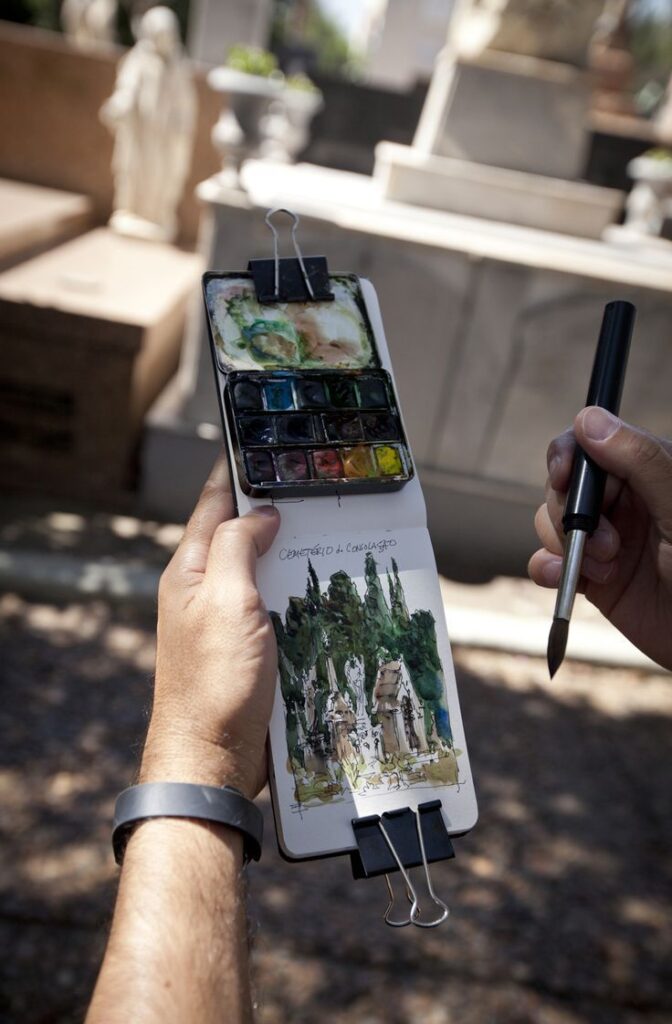 Artist sketching outdoors with watercolor palette, depicting a cemetery scene in a notebook.