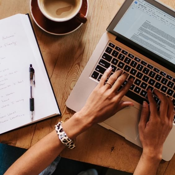 Person typing on a laptop with a notebook and coffee on a wooden table, focused on work.