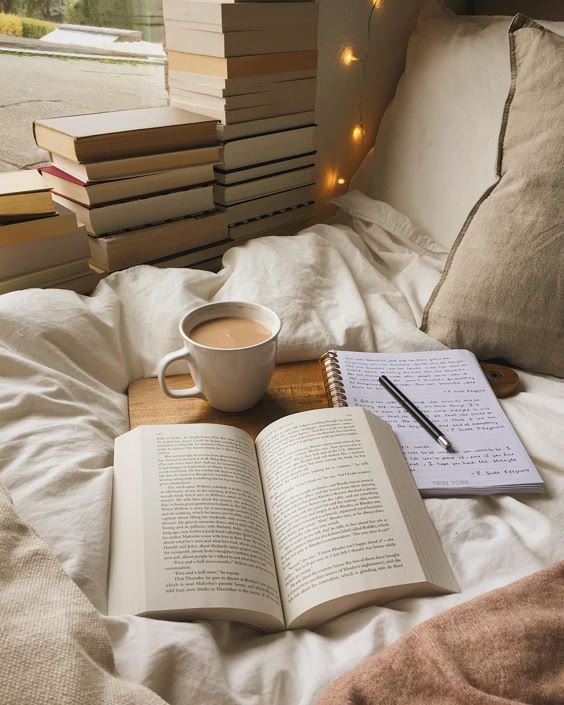 Cozy reading nook with open book, coffee, and notes on a bed, surrounded by stacked books and warm lighting.