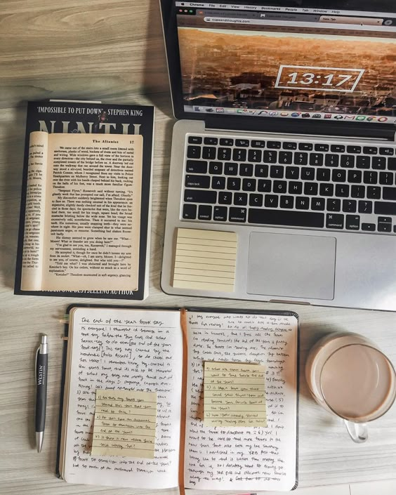 Laptop, open book, journal with notes, and coffee mug on a wooden desk, ideal setting for productive writing or study.