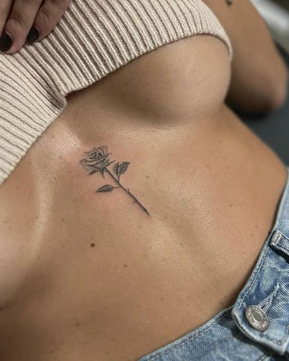 Minimalist rose tattoo under bust on woman's ribcage, wearing a knit top and jeans.