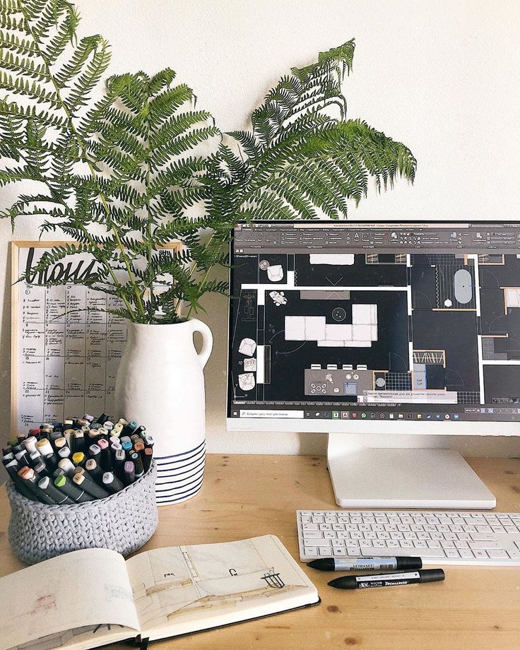 Modern workspace with computer displaying architectural design, surrounded by sketchbook, pens, and a potted plant.