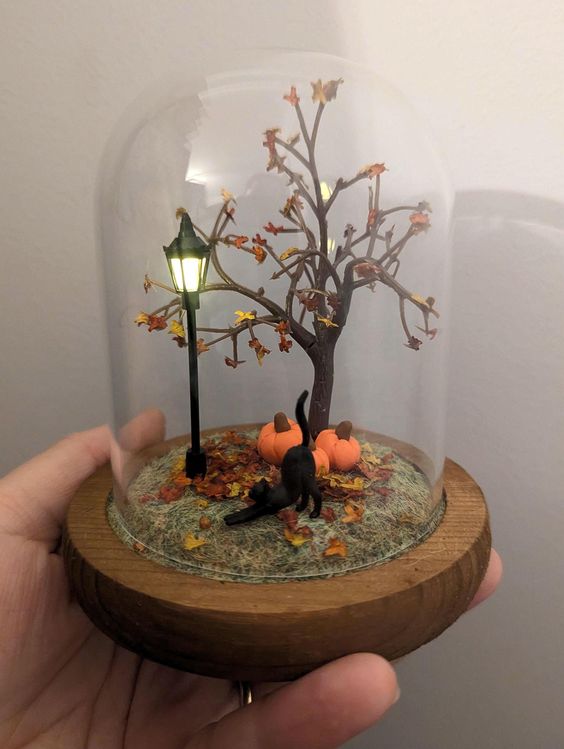 Mini autumn scene with tree, pumpkins, black cat, lamp post inside glass dome. Cozy fall decoration on wooden base.