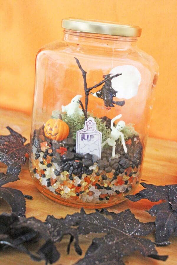 Halloween terrarium in a jar with skeletons, pumpkin, and gravestone on colorful pebbles. Festive decoration.