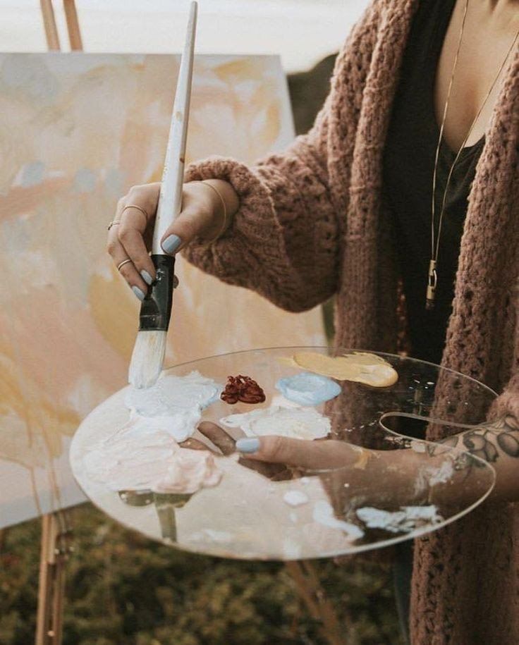 Artist mixing paints on a palette in front of an easel while wearing a cozy sweater, outdoor painting session.