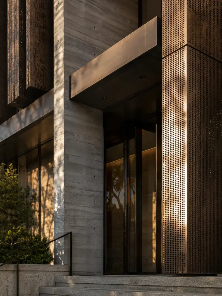 4d6b3bdf-ac23-4395-8af5-014711c775c5 Modern building entrance with textured concrete facade, bronze perforated metal panel, stone steps, warm lighting | Sky Rye Design Modern building entrance with textured concrete facade, bronze perforated metal panel, stone steps, warm lighting