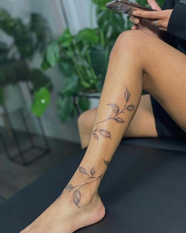 Leg tattoo of delicate leaf vine design on a person sitting near lush green plants.