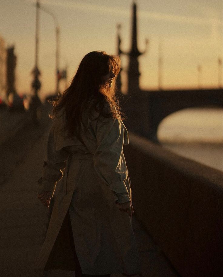 Woman in a trench coat walking on a bridge at sunset, serene and contemplative ambiance.