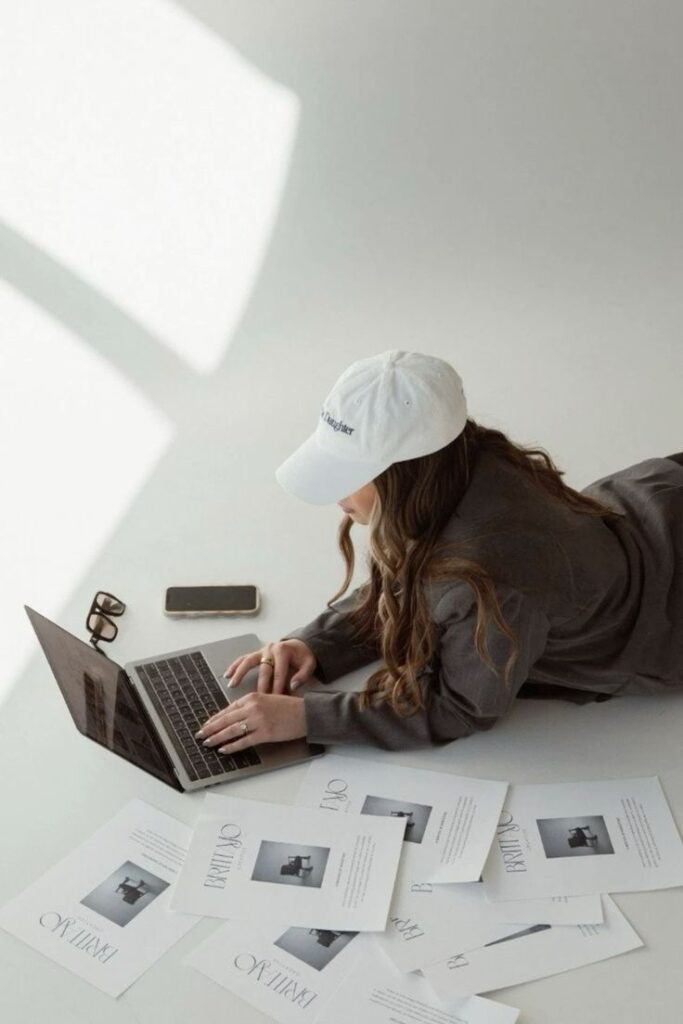 Woman working on laptop surrounded by papers, wearing a white cap, glasses, and smartphone placed nearby.