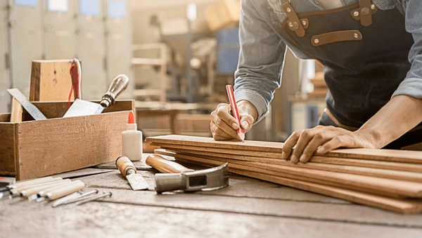 Carpenter marking wood in a workshop, surrounded by tools and wooden planks, in preparation for crafting furniture.