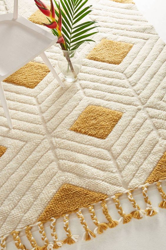 Modern geometric rug with tassels and vase on a white chair, featuring a gold and cream pattern.