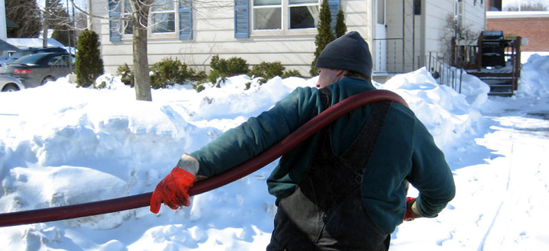 Person in winter clothing carrying a hose outside a snow-covered house.