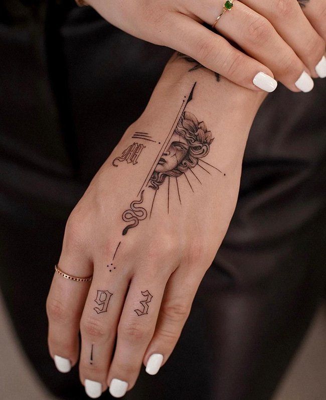 Hand tattoo featuring a Medusa design, the letter M, and numbers 93 on fingers, with minimalistic black ink.