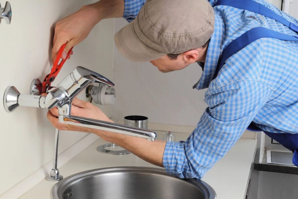 Plumber fixing kitchen faucet with wrench, wearing cap and checkered shirt, close-up on sink and tools.