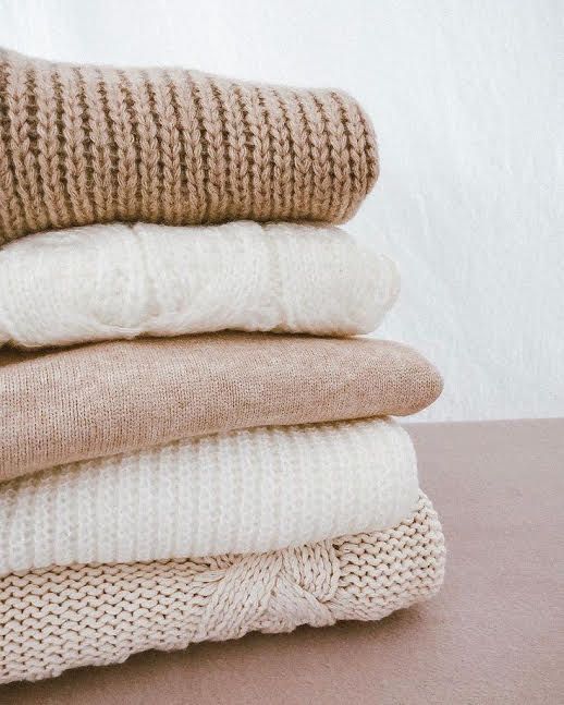 Stack of cozy neutral-colored sweaters folded neatly on a flat surface.