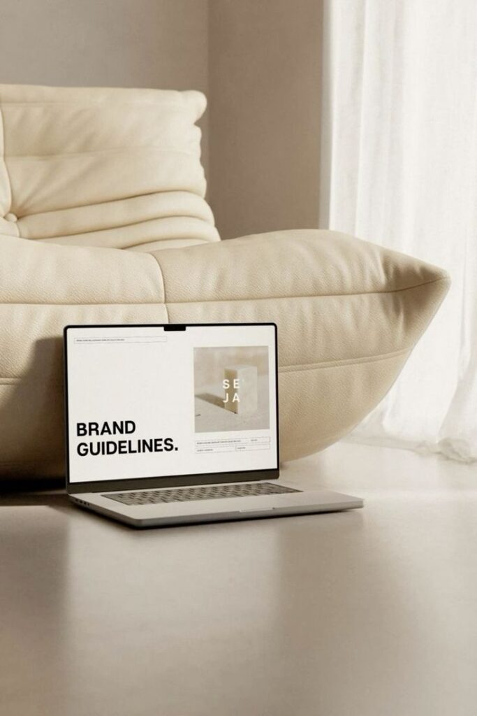 Laptop displaying brand guidelines document on screen, placed on floor beside cozy cream sofa in softly lit room.