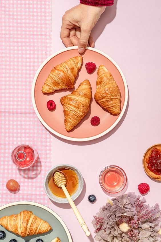 Plate of croissants with raspberries, honey, and juice on a pastel table setting, hand placing the plate.