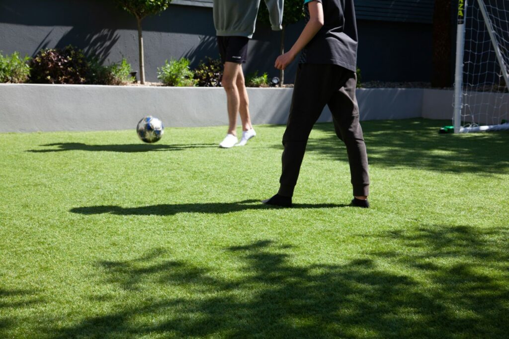 Two people playing soccer on a sunny yard with artificial grass, next to a goalpost and green plants.