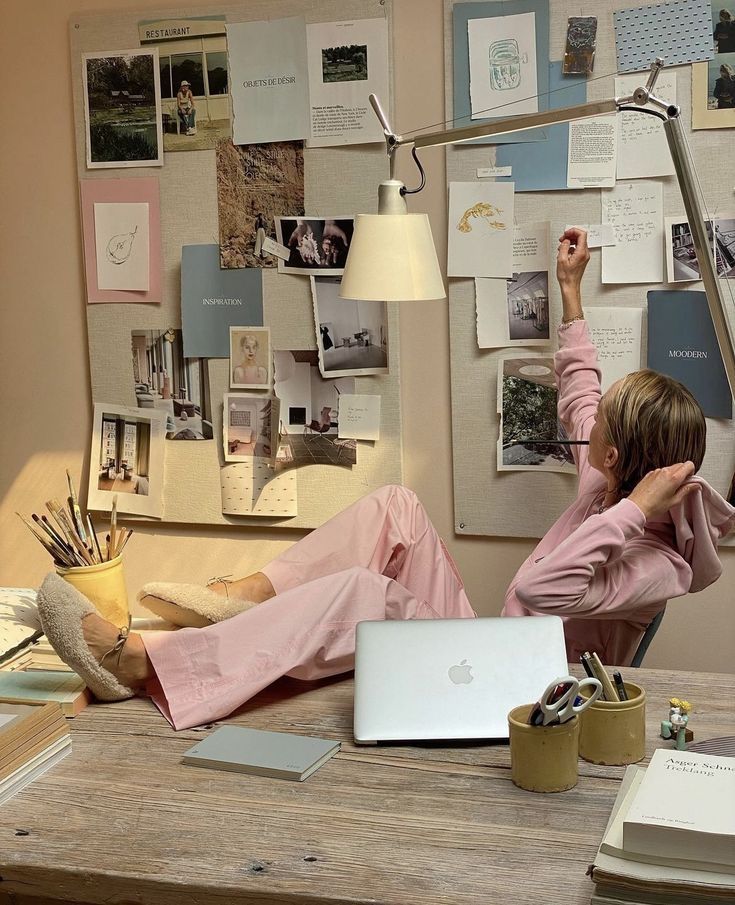 Person in pink leisurewear brainstorming with mood board and laptop, feet up on desk, in cozy creative workspace.