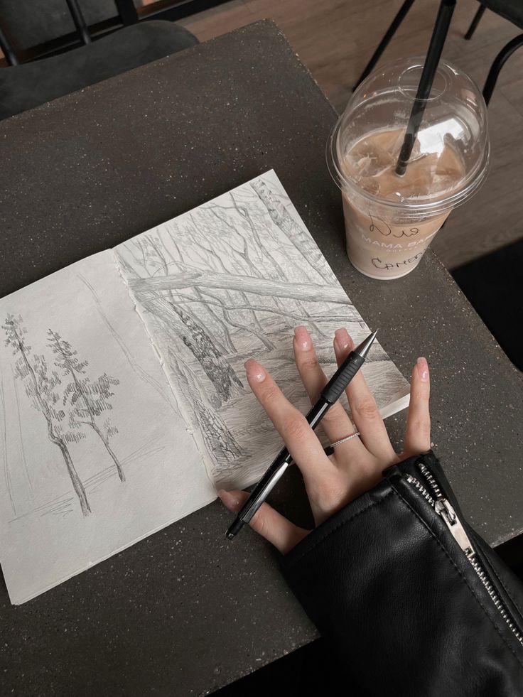 Hand sketching nature scene in notebook at café, with iced coffee and pen on table. Art and relaxation concept.