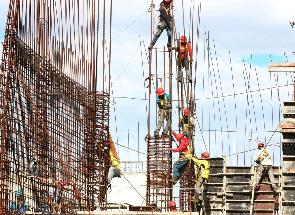 Construction workers on a high-rise project with safety gear, reinforcing steel bars under clear skies.