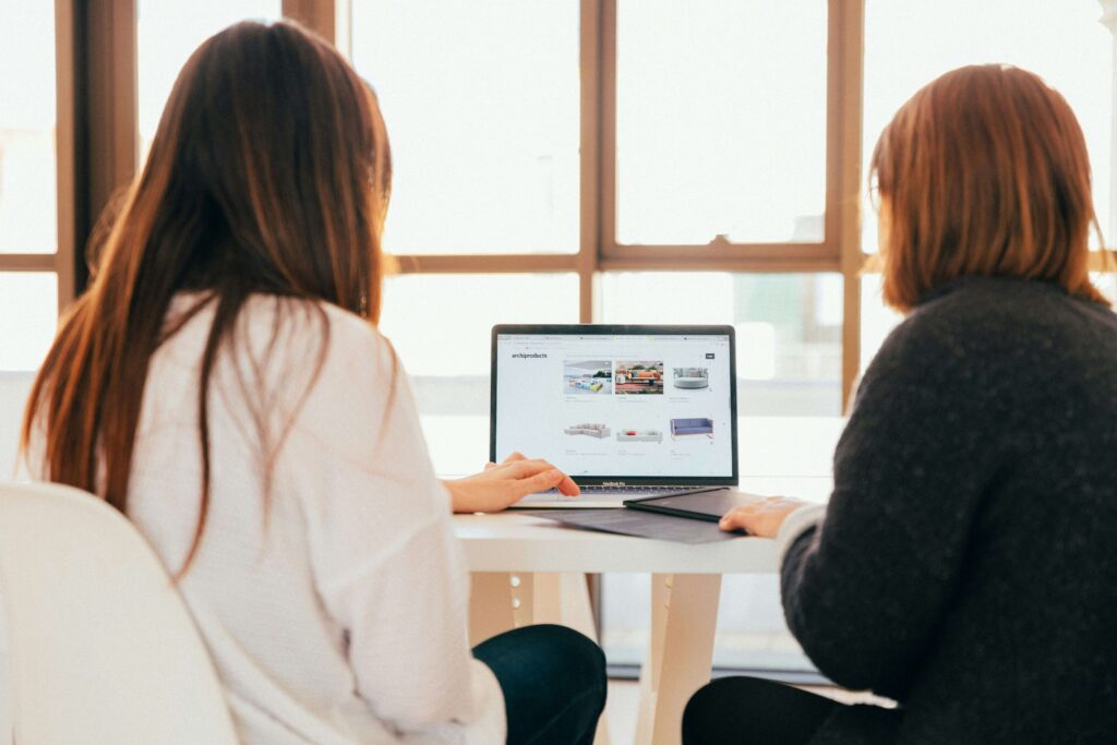 Two women collaborate at a desk, browsing a design website on a laptop, with bright windows in the background.