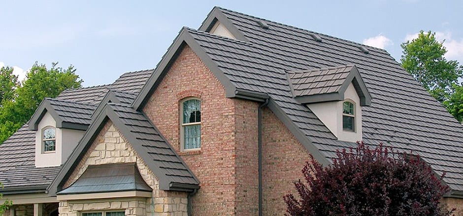 Brick house with gabled roof and gray shingles surrounded by trees under a clear sky.