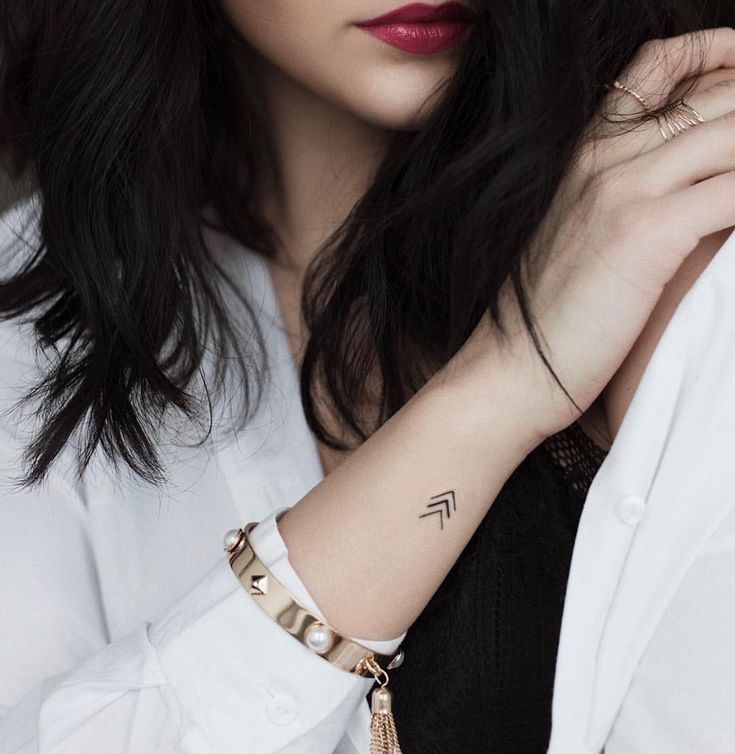 Chic woman with arrow wrist tattoo, gold bracelet, and dark hair in a white shirt.