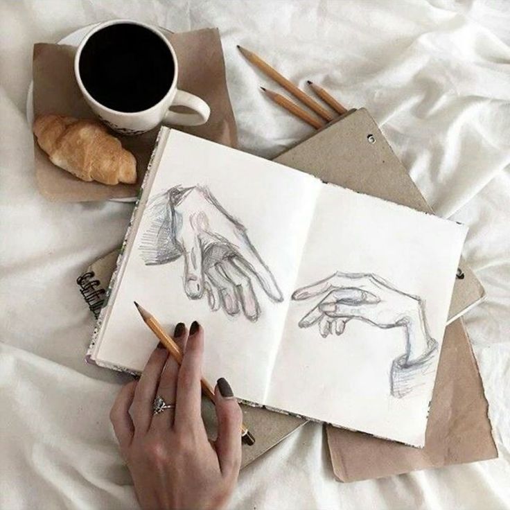 Open sketchbook featuring hand drawings with a coffee and croissant nearby, pencils, and a hand holding a pencil.