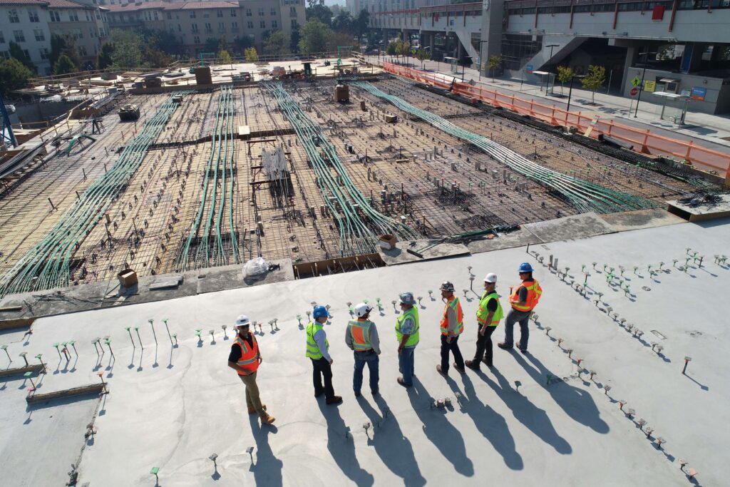 Construction team reviews large building site, focusing on foundation and groundwork under clear sky.