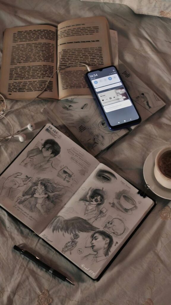 Open book, smartphone, sketchbook with drawings, cup of coffee, and pen on a cozy bed setting in warm lighting.