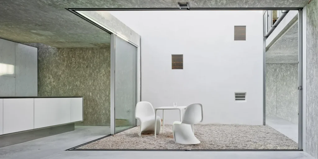 Minimalist indoor patio with white chairs and table on gravel, surrounded by modern concrete walls.