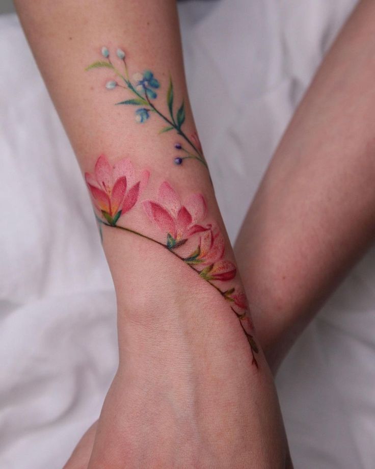 Floral wrist tattoo with pink and blue flowers and green leaves on arm, showcasing delicate, colorful design.