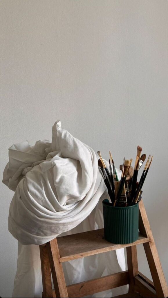 Painting tools on a wooden stool with a white cloth, against a minimalistic background.